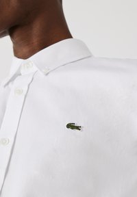 Chemise blanche à col avec une fermeture à bouton, arborant un petit logo de crocodile vert brodé sur le côté gauche de la poitrine.