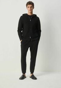 Schwarze Zip-Hoodie mit Kapuze, kombiniert mit schwarzen schmalen Jogginghosen und schwarzen Slippers. Lässiges Design, weicher Stoff, minimale Details.