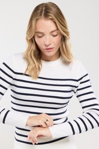 Vrouw met schouderlang blond haar die een witte trui met zwarte horizontale strepen draagt, terwijl ze de mouwen met beide handen aanpast.