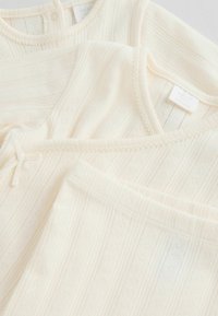 Ensemble de vêtements pour bébé de couleur crème douce avec un motif texturé délicat, comprenant un haut à manches longues, un body et un pantalon avec bordure côtelée.