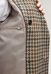 Mantel aus Wollmischung mit Hahnentrittmuster in Erdtönen. Das Futter verfügt über eine Reißverschlusstasche und eine kontrastierende glatte Textur. Schwarzes Knopfdetail.