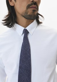 Chemise blanche à col écarté, assortie d'une cravate texturée bleu foncé arborant un motif subtil. Vue en gros plan.