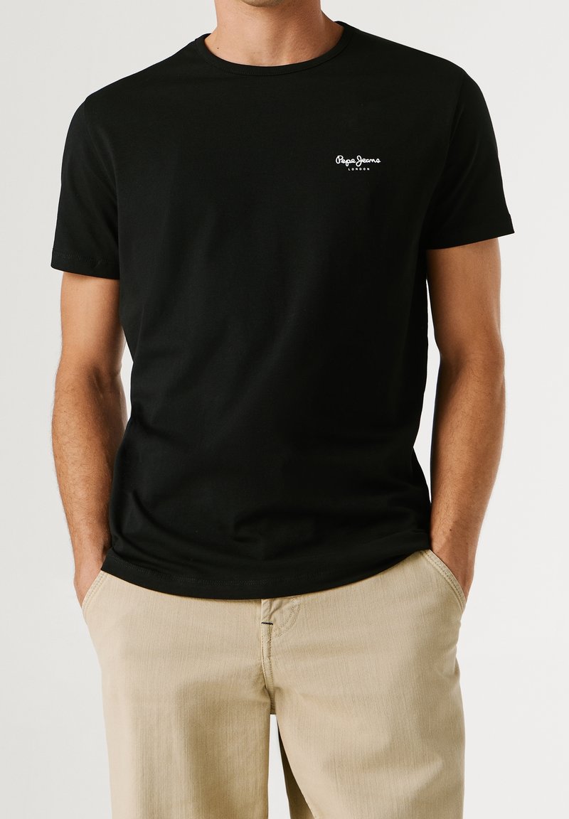 Homme portant un t-shirt noir Pepe Jeans London et un pantalon beige, les mains dans les poches, sur un fond uni.