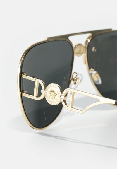 Ochelari de soare negri cu un cadru din metal aurit. Caracteristici notabile includ un medalion decorativ cu un leu și un design distinctiv al punții.