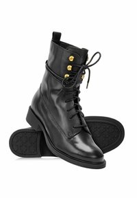 Czarne skórzane botki za kostkę z sznurowanym przodem, złotymi oczkami, pętelką do zakupu i wzorzystą czarną gumową podeszwą, ułożone tak, że jeden but spoczywa na drugim.