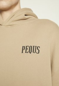 Bežs džemperis ar kapuci, izgatavots no teksturēta auduma, ar izteiktu melnu "PEQUS" logo uz krūšu zonas. Klasisks kapuču dizains ar ribotām akcentiem.