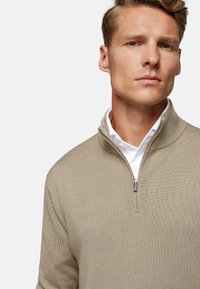 Maglione bebè a mezza zip beige con un motivo a maglia testurizzata. Presenta una chiusura con zip e una camicia bianca sotto.