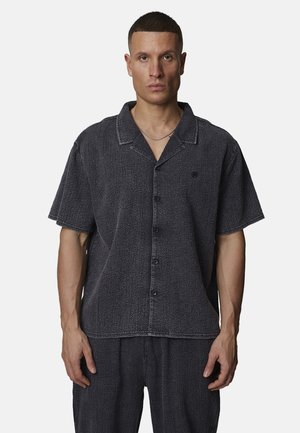 Uomo con camicia a maniche corte grigio scuro con colletto e pantaloni coordinati, in piedi davanti a uno sfondo chiaro e semplice.