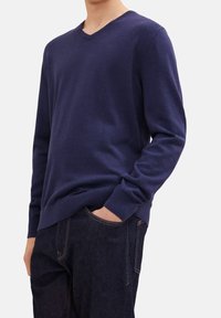 Marineblauer Pullover mit V-Ausschnitt aus weichem Stoff, mit langen Ärmeln und einem gerippten Saum. Kombiniert mit dunklen Denim-Jeans.