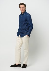 Camicia di denim blu scuro con bottoni davanti, abbinata a pantaloni bianco sporco. Mocassini neri completano l'outfit. Design semplice e classico.