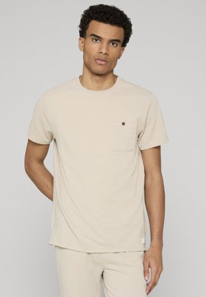 T-shirt beige à manches courtes en tissu texturé, doté d'un col rond et d'une poche poitrine avec un petit bouton noir.