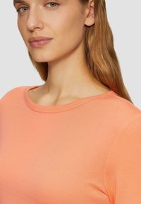 Licht perzikkleurig t-shirt met een ronde hals, korte mouwen en een zachte, aansluitende textuur. Sichtbare stiksels langs de halslijn.
