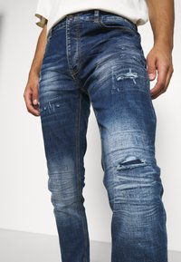 Tmavě modré džínové kalhoty s úzkým střihem, s poškozenými místy a vyblednutím. Žluté prošívání podél švů dodává kontrast.