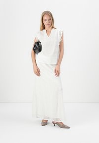 Weiße Rüschenshirt mit V-Ausschnitt und Brokatdetails, kombiniert mit einem langen weißen Rock mit Spitzenmustern und grauen spitzen High Heels.