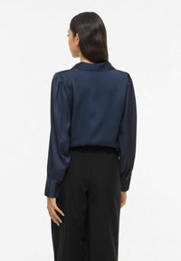 Neredzas zila satīna blūze ar klasisku apkakli, garām pūrstām piedurknēm un pogām aizpogājamām aprocēm, kombinēta ar melnām piegrieztām biksēm. Gluda tekstūra.