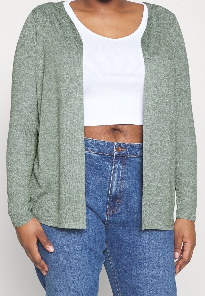 Groene gebreide cardigan met open voorkant, lange mouwen en een zachte textuur, gecombineerd met een witte cropped tanktop en denim jeans.