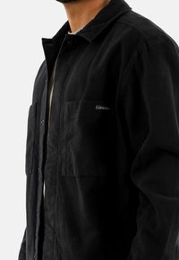 Chaqueta negra hecha de tela texturizada, con cuello de camisa, botones frontales y dos bolsillos en el pecho; marcada con un logo de Calvin Klein.