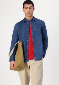 Denim knapp-skjorte i mørkeblå, rød t-skjorte under, lyse beige bukser, holder en lys beigefarget kanvas tote bag, med detaljer i brystlomma.