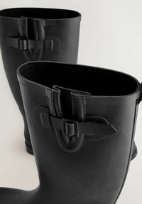 Des bottes en caoutchouc noires avec un haut large et arrondi, ornées de sangles décoratives avec des boucles. L'intérieur est texturé, offrant adhérence et confort.