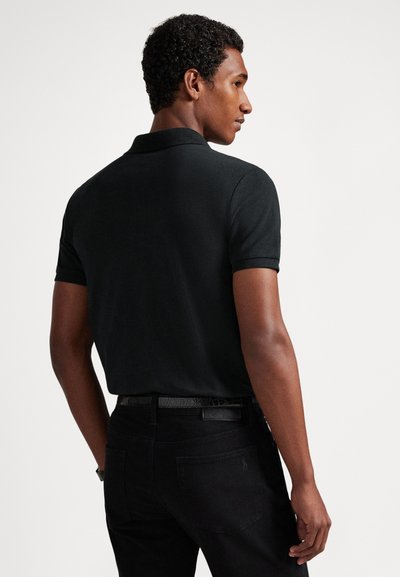 Melnā īsroku polokrekls no gludas auduma ar klasisku apkakli un piegulošu dizainu. Valkāts kopā ar melnām biksēm un teksturētu jostu.