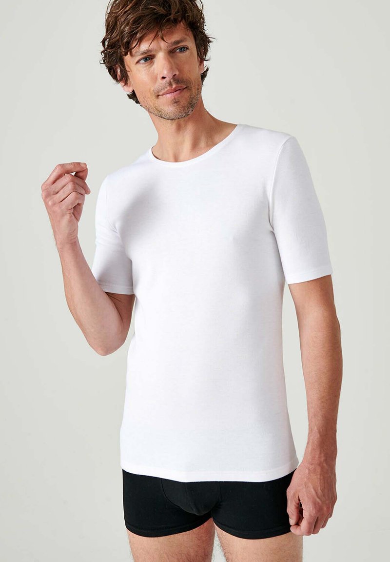 T-shirt bianca a maniche corte realizzata in tessuto liscio, con scollatura rotonda e design aderente. Indossata con pantaloni corti neri.