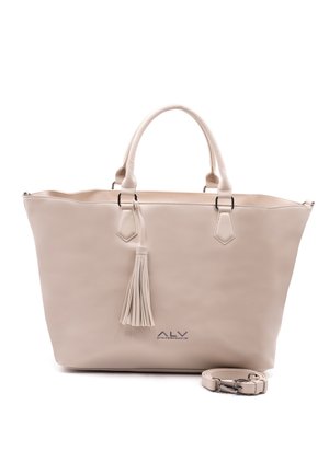 Sac fourre-tout beige avec anses doubles, un pompon décoratif et une bandoulière amovible enroulée à côté sur fond blanc.