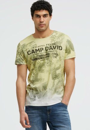 Mann mit lockigem Haar trägt ein hellgrünes und weißes grafisches T-Shirt und blaue Jeans und steht mit einer Hand in der Tasche vor einem schlichten Hintergrund.