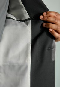 Interior de una chaqueta de traje negra con un forro suave de color gris claro. Un bolsillo único en el lado izquierdo y un borde texturizado negro a lo largo del borde.