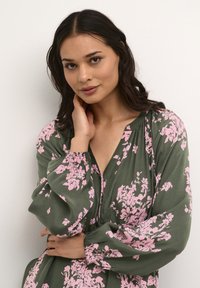 Rochie florală verde cu modele roz, mâneci lungi, decolteu în V și detalii pliate la umeri. Material ușor, croi lejer.