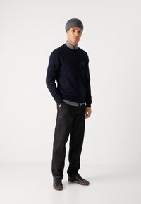 Navy knit sweater with a round neckline, gray collared shirt underneath, dark denim pants, and gray knit beanie. Casual footwear in black.