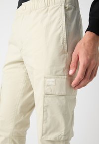 Helle beigen Cargo-Hosen aus einem strukturierten Stoff. Mit einem elastischen Bund, seitlichen Taschen und einem Logo-Patch am Oberschenkel.