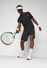 Zwarte katoenen T-shirt met een klein groen logo, gecombineerd met grijze shorts en witte sportschoenen; houdt een tennisracket vast met een gele bal.