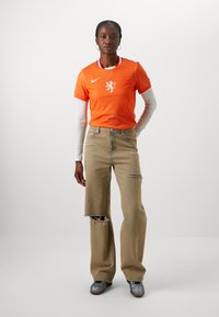 Orange Nike-tröja med ett lejonlogo och prickig textur, kombinerad med bruna slitna vida jeans och grå skor.