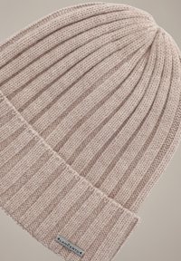 Jasnoróżowa dziana czapka z pionowym prążkowaniem, z podwiniętym mankietem i małą metalową metką. Miękka tekstura i zaokrąglony kształt.