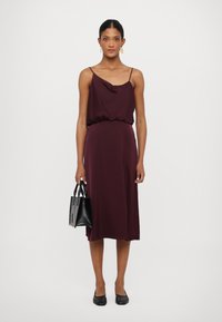 Rochie midi bordeaux cu guler drapat, talie elastică și bretele subțiri. Împreună cu o poșetă mică neagră și balerini negri.