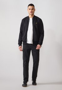 Juodas zip-up bomber tipo švarkas su gumuotomis rankovėmis ir maža geltona logo, dėvimas ant balto marškinėlio ir juodų laisvalaikio kelnių, derinamas su juodais sportbačiais.