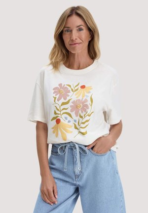 Vrouw draagt een wit T-shirt met een roze en gele bloemenprint, ingestoken in lichtblauwe denim jeans met een strikceintuur.