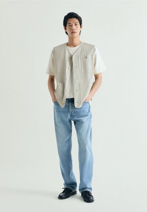 Jonge man met een beige vest met knopen over een wit T-shirt, lichtblauwe spijkerbroek en zwarte schoenen, staand met handen in de zakken tegen een effen witte achtergrond.