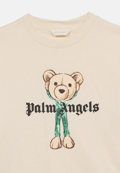 Μπεζ μπλουζάκι με γραφικό που απεικονίζει ένα άτομο με μεγάλη κεφαλή αρκούδας τεντ και πράσινη ενδυμασία, κείμενο «Palm Angels» σε μαύρη γοτθική γραμματοσειρά.