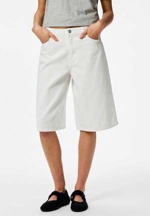 Personne portant un short blanc ample mi-cuisse, un haut gris court et des chaussures plates noires, les mains dans les poches, debout sur un fond blanc.