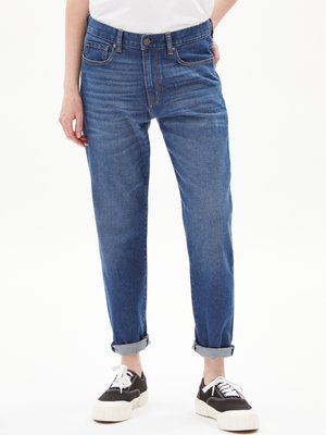 Persona che indossa jeans blu arrotolati e sneakers nere con lacci bianchi, in piedi contro uno sfondo bianco uniforme.