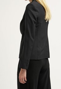 Blazer noir ajusté avec manches longues, coupe cintrée et épaules légèrement rembourrées. Détails remarquables : boutons décoratifs aux poignets.