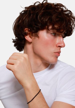 Black string bracelet on a light-skinned male's wrist, wearing a white T-shirt, with curly brown hair and a side profile.