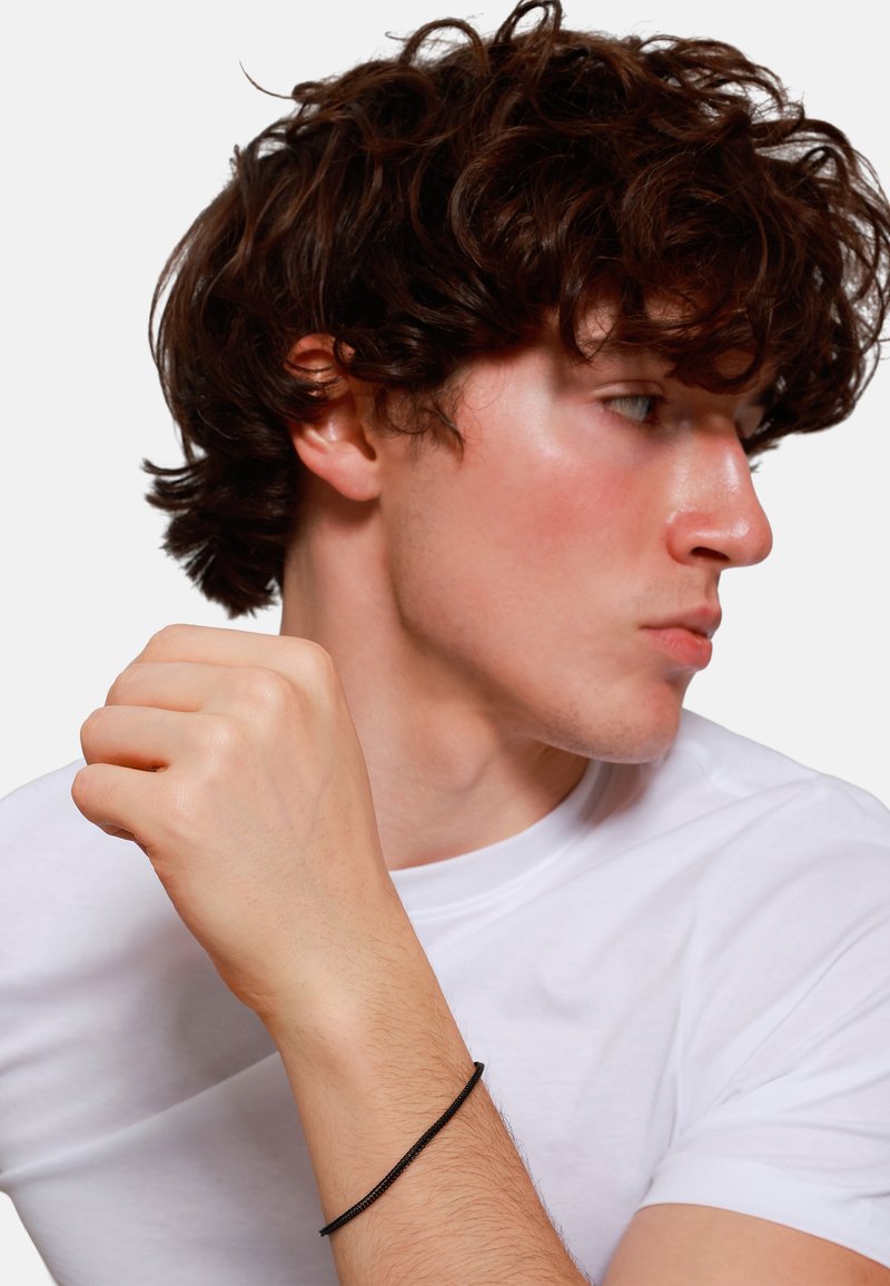 Svart trådarmband på en lyst skinnet manns håndledd, iført en hvit T-skjorte, med krøllete brunt hår og sideprofil.