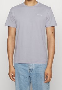 Camiseta de algodón de color púrpura claro con mangas cortas, escote redondo y pequeño logo blanco de "Calvin Klein" en el lado izquierdo del pecho.