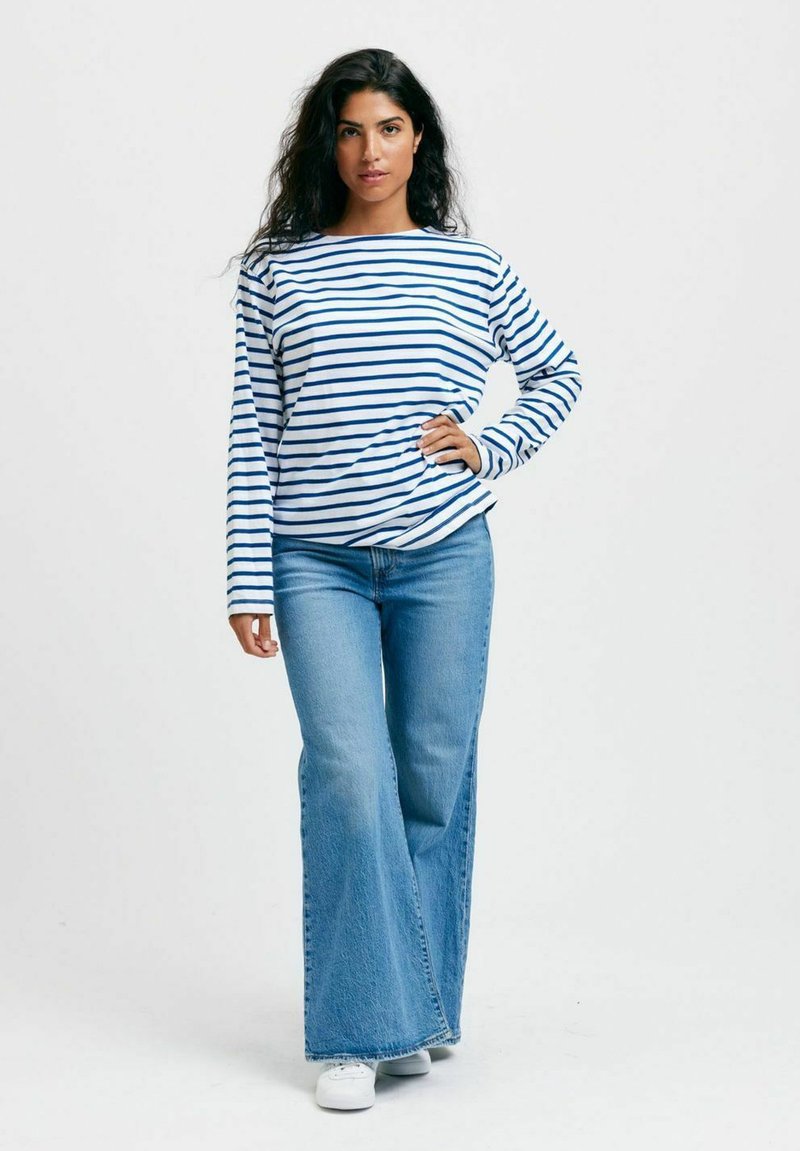 Pitkähihaisen, tummansinisen ja valkoisen raidallisen paidan parina on leveälahkeiset, vaaleansiniset farkut ja valkoiset kengät. Yksinkertaista, rentoa pukeutumista, jossa on väljä kaarroke.