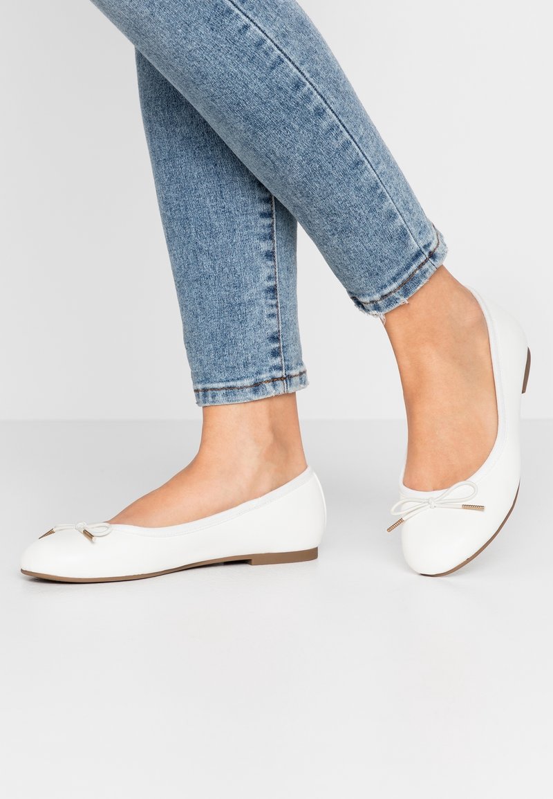 Pieds en ballerines blanches avec petits nœuds portées avec un jean bleu clair court sur un fond gris uni.