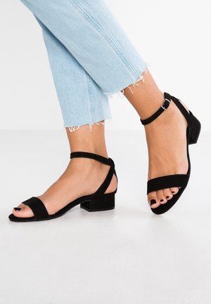 Füße in schwarzen Wildleder-Sandalen mit Knöchelriemen und Blockabsätzen, kombiniert mit hellblauen, ausgeblichenen Jeans mit cropped und fransigem Saum.