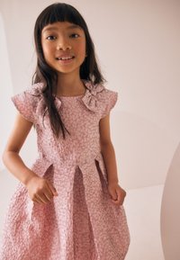 Hele-roosa kleit tekstuurse kangaga, millel on kapuutsivarukad ja õlgadel lipsudetailid, lõikega, mis sisaldab voltidega seelikut.