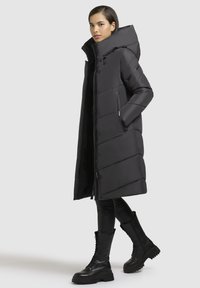 Must mustri jakkk ja kõrge krae ja kapuutsiga, vertikaalse tikkimisega, lukuga sulgemine ja esikotid. Kantud läikivate mustade retuuside ja saapatega.